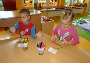 Dwoje dzieci siedzi przy stoliku, w ręku trzyma kredki i układa je na stole według wzoru na kartkach.